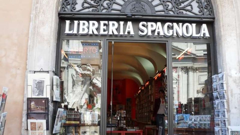 Ingresso di una libreria con insegna “LIBRERIA SPAGNOLA”, incorniciato da un grande arco in pietra e da una lunetta in ferro battuto decorata. Vetrine laterali con libri e cartoline esposte e porta a vetri che lascia intravedere l’interno con soffitto a volte e scaffali.