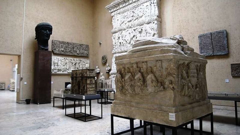 Sala museale con pareti beige e pavimento in pietra, con grandi reperti archeologici esposti su supporti neri, tra cui un sarcofago scolpito in primo piano. Alle pareti sono presenti rilievi e lastre decorative, oltre a un grande busto scuro su piedistallo.