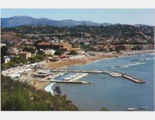 Veduta panoramica di una cittadina costiera con lunga spiaggia attrezzata e un porto turistico con moli e barche. Sullo sfondo si vedono edifici e colline verdeggianti che salgono verso montagne lontane.