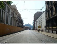 Strada urbana ampia tra edifici alti, con binari del tram sulla carreggiata e cavi aerei sospesi. Ai lati si vedono facciate storiche e moderne, un lungo cantiere recintato e marciapiedi con segnaletica e lampioni.