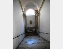Scala interna in pietra che scende verso un piccolo pianerottolo, con pareti bianche e corrimano in ferro ai lati. In fondo si vede un arco con finestra semicircolare e decorazioni architettoniche in pietra, con un busto su mensola sulla parete.