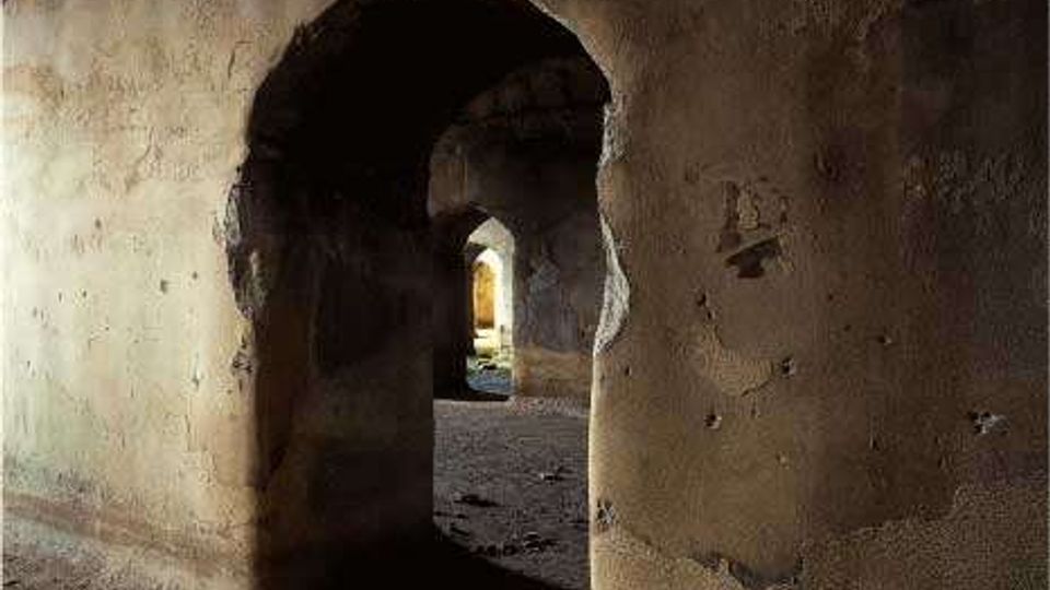 Interno di un edificio antico in muratura con intonaco consumato, caratterizzato da archi in sequenza e pareti spesse. Un corridoio scuro conduce verso un’apertura luminosa in fondo, con pavimento in terra e pietrisco.
