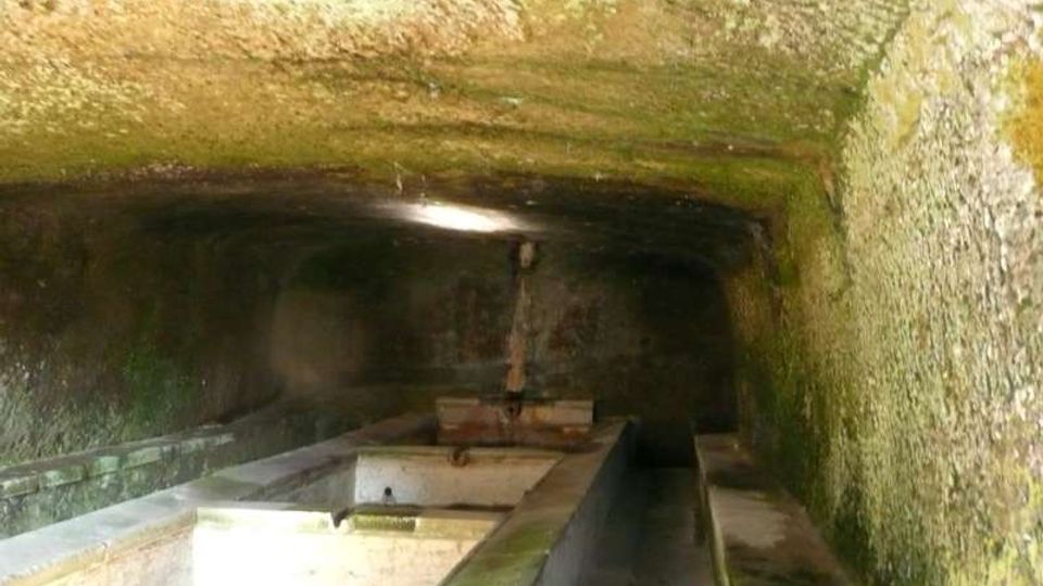 Interno di una vasca coperta in pietra, con soffitto basso e pareti umide ricoperte di muschio. Al centro corre un lungo canale in muratura con comparti rettangolari, illuminato debolmente da una piccola apertura superiore.