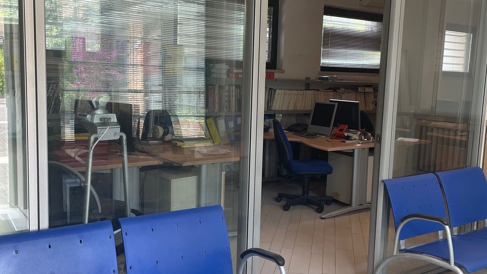 Sala d’attesa in un ufficio, con sedute blu in plastica su pavimento chiaro e pareti a vetrate con telai metallici. Dietro le vetrate si vedono stanze con scrivanie, computer, scaffali di documenti e finestre con veneziane.