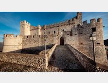 Castello medievale in pietra con mura merlate e torri, visto dall’esterno. Un ponte in pietra conduce all’ingresso ad arco, con lampioni lungo il percorso e cielo sereno sullo sfondo.