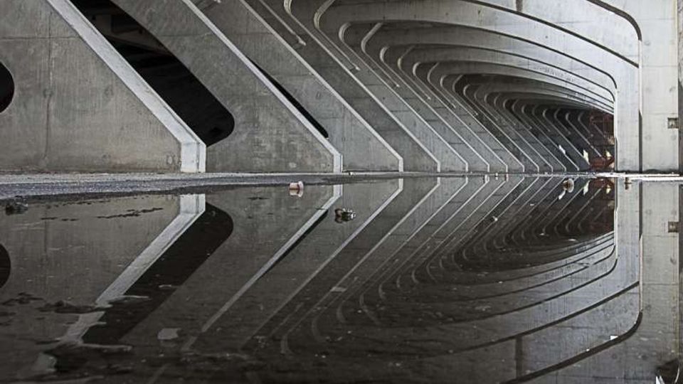 Sottopasso in cemento con una serie di pilastri e archi curvi ripetuti lungo il soffitto, in prospettiva. Una grande pozzanghera in primo piano riflette la struttura e le linee dell’architettura.