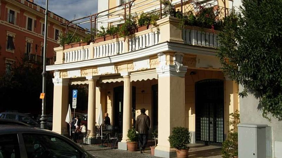 Edificio ad angolo in stile classico con portico a colonne e balconata superiore, con ringhiera e fioriere su una terrazza. Davanti si vede un marciapiede in pietra con arredi esterni e piante in vaso, affacciato su una strada urbana con lampione e auto parcheggiate.