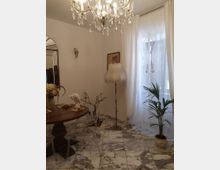 Interno di una sala da pranzo con pavimento in marmo, tavolo rotondo in legno e sedie trasparenti, illuminata da un lampadario di cristallo. Accanto a una grande finestra con tende bianche ci sono una pianta in vaso e una lampada da terra, con specchio e applique a parete.