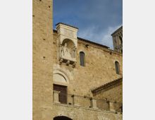 Facciata esterna di un edificio storico in pietra, con finestre ad arco e un portale sormontato da una nicchia sporgente con scultura e baldacchino decorato. In primo piano si vede una terrazza con ringhiera in ferro e, sullo sfondo, un tratto di torre e cielo.