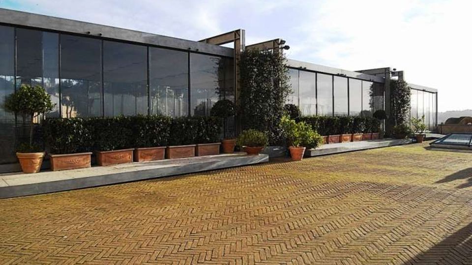 Terrazza esterna pavimentata in mattoni a spina di pesce, con una lunga facciata moderna in vetro e struttura metallica. Lungo il perimetro ci sono fioriere e vasi con siepi e alberelli, sotto un cielo aperto.