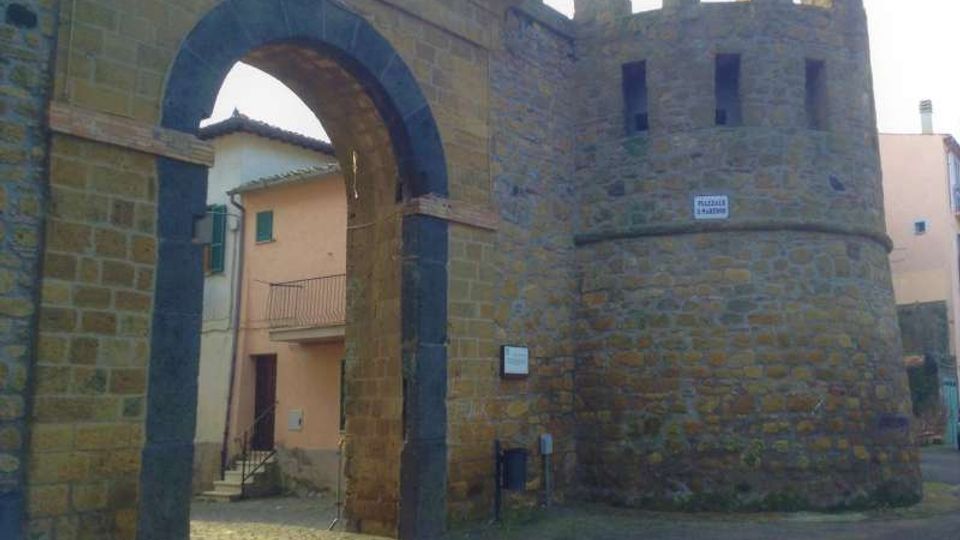 Antica porta urbana in pietra con grande arco d’ingresso e torre cilindrica merlata adiacente. Sullo sfondo si vedono edifici residenziali e una strada lastricata che attraversa il varco.