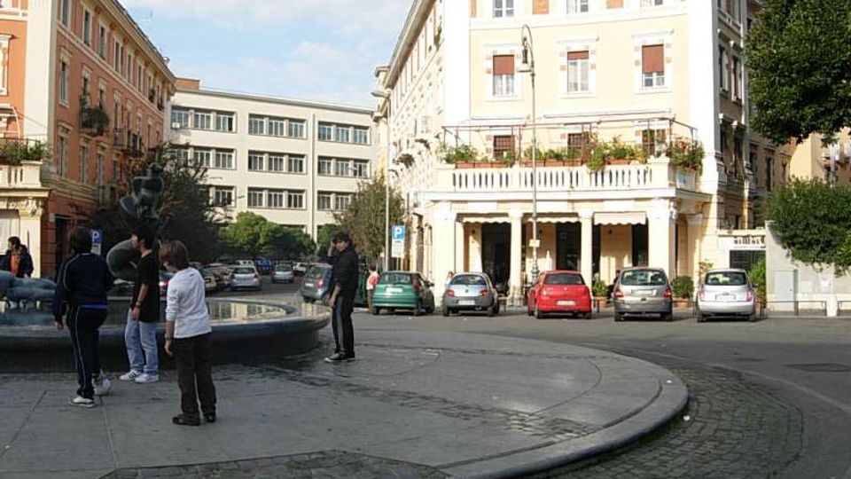 Piazza urbana con una fontana circolare in primo piano e una strada curva lastricata che la costeggia. Sullo sfondo si vedono palazzi a più piani con facciate chiare e balconi, auto parcheggiate lungo il bordo e alberi sul lato destro.
