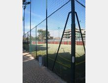 Campo sportivo all’aperto con superficie in erba sintetica e linee gialle, circondato da alte recinzioni in rete metallica e illuminazione su pali. In primo piano si vede un marciapiede in pietra lungo la recinzione, con edifici residenziali sullo sfondo.