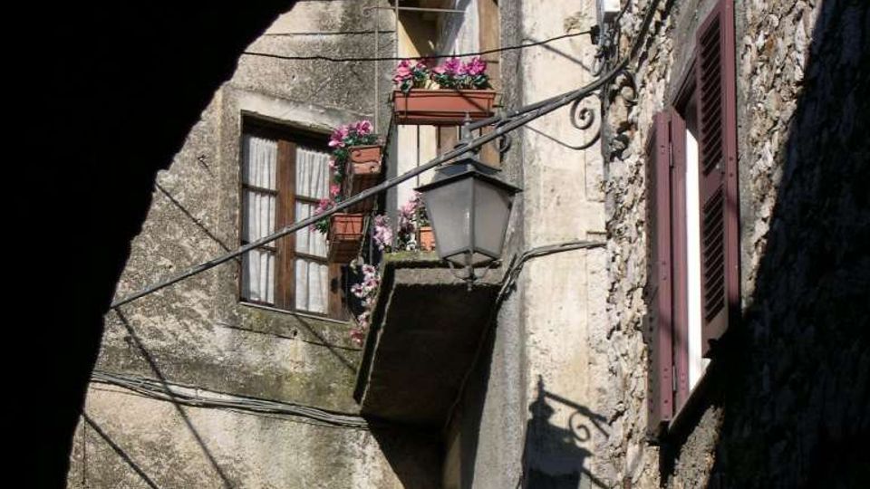 Facciate di case in pietra e intonaco in un vicolo stretto, con balconcino decorato da fiori, lampione a parete e cavi elettrici a vista. Finestre con persiane scure e muri irregolari creano un ambiente urbano storico.