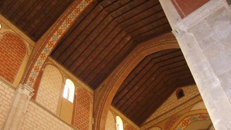 Interno di una chiesa con soffitto a capriate in legno scuro e grandi archi in mattoni decorati con motivi geometrici. Pareti in pietra chiara con finestre ad arco che lasciano entrare luce naturale.
