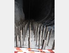 Interno di un grande tunnel o condotta scura, con pavimento in calcestruzzo e file fitte di pali verticali arrugginiti disposti in corridoi. In primo piano si vede una barriera a nastro bianco e rosso che delimita l’area.