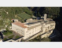 Veduta aerea di un grande complesso monastico in pietra con tetti in coppi, cortili interni e un alto campanile quadrato. L’edificio è affacciato su una scarpata e circondato da colline boscose, campi coltivati e un piccolo parcheggio.