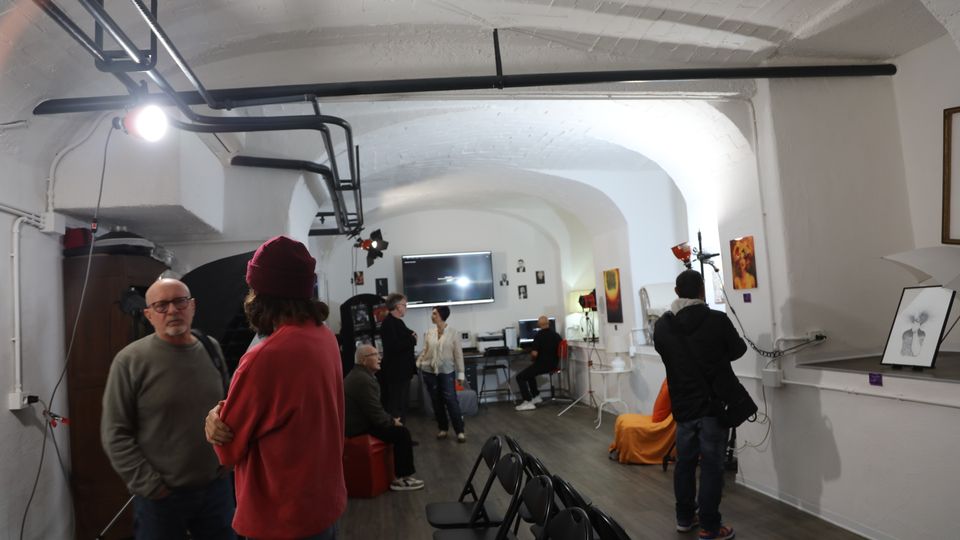 Interno di uno spazio tipo studio o piccola galleria con soffitto a volta in mattoni dipinti di bianco e tubazioni a vista, illuminato da luci da set. Lungo la sala ci sono sedie pieghevoli, attrezzatura video e fotografica, schermi e quadri appesi alle pareti, con pavimento in legno scuro.