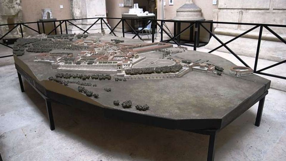Grande plastico architettonico di una città antica esposto su un tavolo poligonale, circondato da barriere nere. L’ambiente è una sala museale con pareti in pietra e archi, con elementi scultorei sullo sfondo.