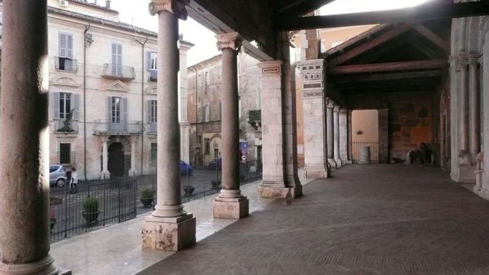 Portico storico con pavimentazione in pietra e lunga fila di colonne in muratura che sorreggono un soffitto con travi a vista. Sul lato aperto si affaccia su una strada e su edifici d’epoca con finestre e balconcini, separati da una ringhiera.