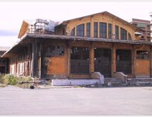 Edificio industriale in mattoni con grande tetto a capanna e ampie finestre ad arco, con portoni metallici e una veranda coperta su pilastri lungo la facciata. Area esterna asfaltata e spoglia, con segni di degrado e strutture metalliche visibili sul lato.