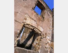 Rovine di un edificio in muratura con pareti intonacate e parti crollate, che lasciano visibili archi e aperture senza infissi. Si notano mattoni e pietre a vista, con il cielo azzurro sullo sfondo attraverso le finestre.