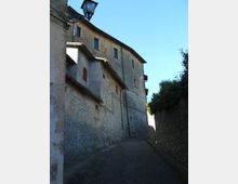 Vicolo stretto e in pendenza tra edifici in pietra e intonaco con piccole finestre e tetti a falda, illuminato da un lampione a muro. A destra corre un muretto con vegetazione, sotto un cielo sereno.