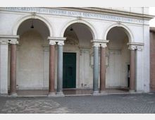 Portico in pietra con tre grandi archi a tutto sesto sostenuti da colonne in marmo di colori diversi, con iscrizione sulla trabeazione e lampade sospese. Al centro si vede una grande porta verde incorniciata da decorazioni, affacciata su un cortile pavimentato in pietra.
