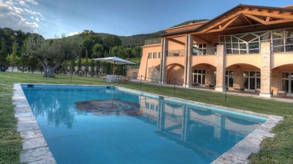 Piscina all’aperto con bordo in pietra su prato, davanti a una grande villa color ocra con portico ad archi, pilastri e tetto in legno a vista. Sullo sfondo si vedono alberi e colline verdi sotto un cielo sereno.