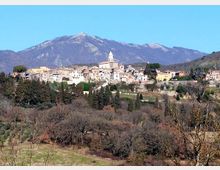 Panorama di un borgo collinare con case in pietra e una chiesa con campanile al centro, circondato da alberi e campi. Sullo sfondo si vede una grande montagna sotto un cielo sereno.