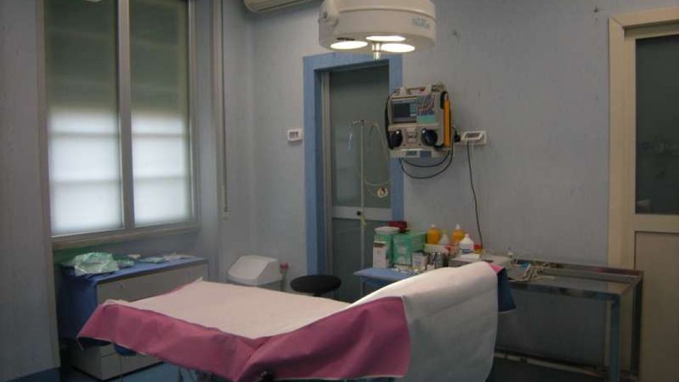 Sala visite medica con lettino coperto da teli bianchi e rosa, lampada chirurgica a soffitto e carrello con strumenti e materiali sanitari. Pareti azzurre con finestra, condizionatore e apparecchiature di monitoraggio fissate al muro.