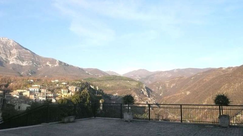 Ampia terrazza panoramica lastricata con ringhiera in ferro e grandi vasi, affacciata su una valle montana. Sullo sfondo si vedono colline e montagne e un borgo con case addossate al pendio sotto un cielo sereno.
