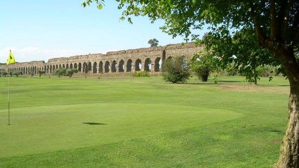 Campo da golf con ampio prato verde e bandierina sulla buca, con un lungo acquedotto in pietra a arcate sullo sfondo. Un albero in primo piano incornicia la scena sotto un cielo sereno.