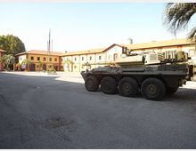 Piazzale asfaltato di una caserma con edifici bassi color ocra e tetti a falde sullo sfondo, con pennoni delle bandiere e palme. In primo piano è parcheggiato un veicolo militare blindato a otto ruote.