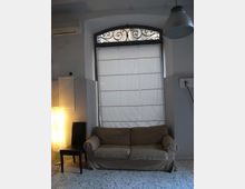 Interno di una stanza con grande finestra ad arco dotata di inferriata decorativa e tenda a rullo bianca. Sotto la finestra c’è un divano a due posti, con una sedia e una lampada da terra su pavimento in graniglia.