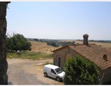 Casale rurale in mattoni con tetto in coppi e camino, affacciato su una strada sterrata con un furgone parcheggiato. Sullo sfondo si estendono campi coltivati ondulati e alberi sotto un cielo sereno.