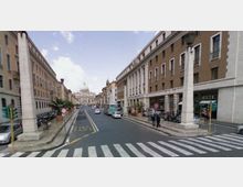 Strada urbana ampia con attraversamento pedonale in primo piano, fiancheggiata da alti edifici in muratura e porticati, con grandi pilastri bianchi e lampioni ai lati. In lontananza si vede la cupola e la facciata della Basilica di San Pietro, con veicoli e segnaletica stradale lungo la carreggiata.