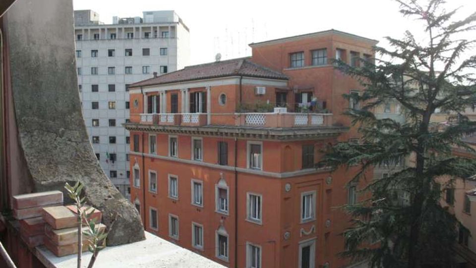 Veduta urbana dall’alto con un edificio in mattoni rossi a più piani, con finestre regolari e terrazze sul tetto con balaustre bianche. Sullo sfondo si vede un alto palazzo bianco e, sul lato destro, un grande albero sempreverde vicino agli edifici.