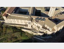 Veduta aerea di un grande complesso storico in pietra con tetti in tegole, cortili interni e un’alta torre campanaria. L’edificio si affaccia su un pendio con mura di contenimento e vegetazione alla base.