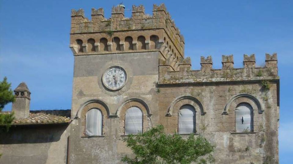 Edificio storico in pietra con torre merlata e orologio circolare sulla facciata, affiancato da un corpo con finestre ad arco e parapetto merlato. Sullo sfondo cielo azzurro, con tetti in coppi e vegetazione in primo piano.