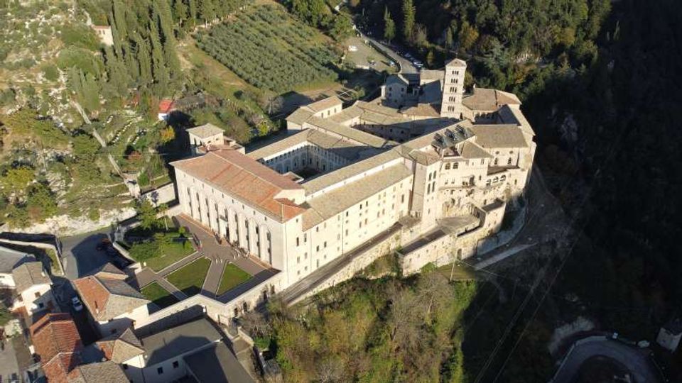 Veduta aerea di un grande complesso monastico in pietra con tetti in coppi, chiostro interno e torre campanaria, affacciato su un pendio. Intorno si vedono strade, case del borgo e campi coltivati con alberi in un’area collinare boscosa.