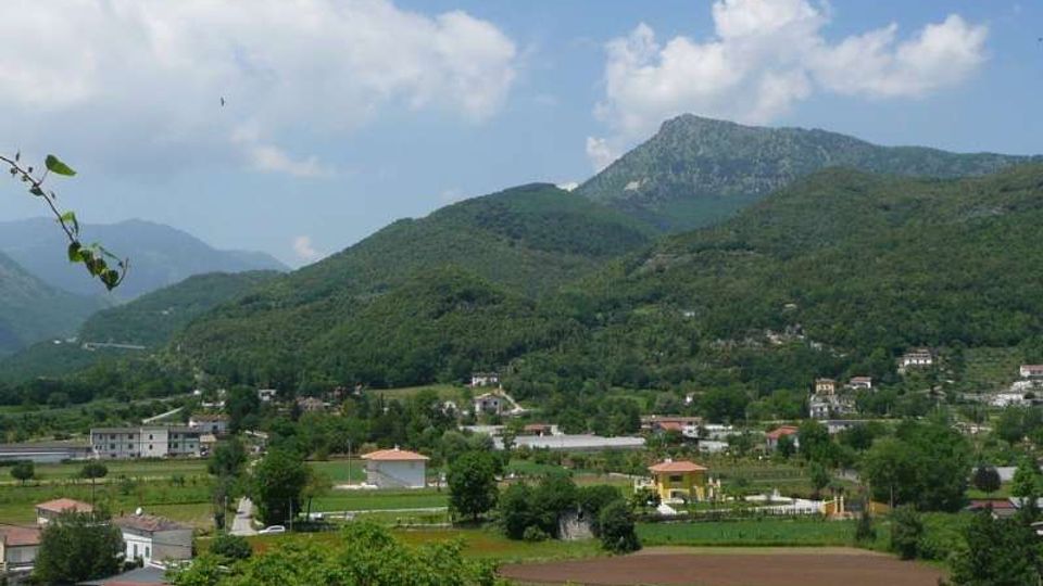 Veduta panoramica di una valle rurale con campi coltivati e case sparse, ai piedi di colline boscose. Sullo sfondo si elevano montagne verdi sotto un cielo azzurro con nuvole.