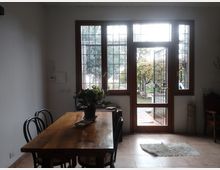 Interno di una sala da pranzo con tavolo in legno e sedie scure, accanto a grandi finestre e a una porta vetrata con inferriata che si apre su un patio e un giardino. Pareti bianche, pavimento piastrellato e tappeto chiaro vicino all’ingresso.