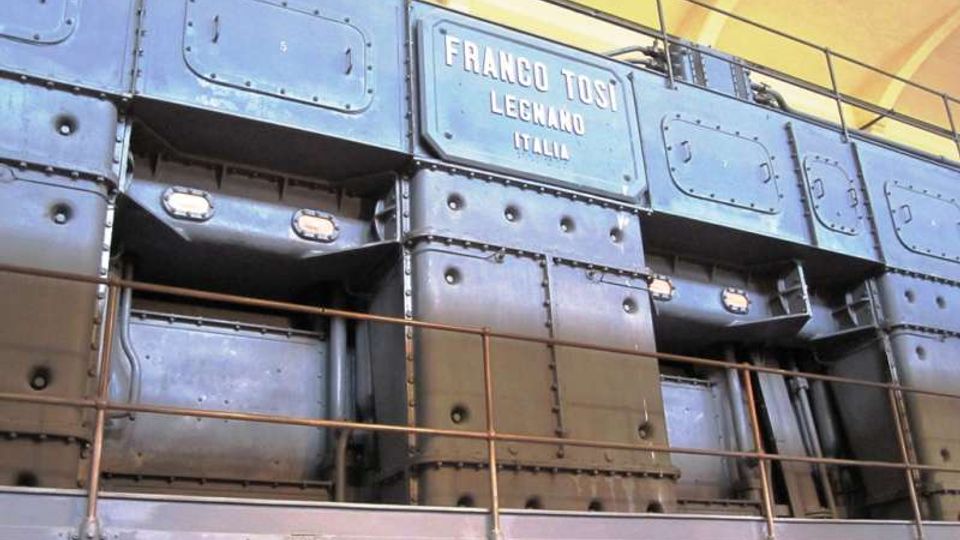 Interno industriale con grandi pannelli metallici rivettati e passerella con ringhiera in metallo, illuminato da luci incassate. Al centro si vede una targa con la scritta “FRANCO TOSI, LEGNANO, ITALIA” sotto una copertura ad arco.