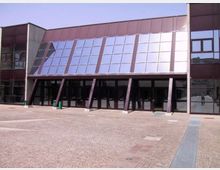 Facciata di un edificio moderno con ampia superficie vetrata inclinata e pannelli metallici, sostenuta da pilastri obliqui sopra un ingresso con porte scure. Davanti si apre una grande piazza pavimentata in pietra, con cielo sereno sullo sfondo.