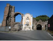 Fontana monumentale addossata a un’antica struttura in mattoni con grandi archi e tratti in rovina, lungo una strada asfaltata. Sullo sfondo si vedono un passaggio ad arco che attraversa il muro e vegetazione in cima alle mura sotto un cielo sereno.