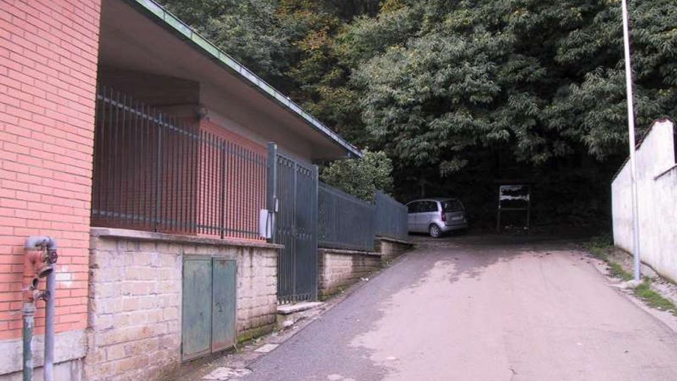 Strada asfaltata in salita che costeggia un edificio in mattoni con recinzione metallica e piccolo accesso, affiancata da un muro e un lampione. In fondo si intravede un’auto parcheggiata davanti a un’area alberata fitta.