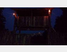 Capanna sopraelevata in legno con parapetto e tetto sporgente, illuminata da due fiaccole ai lati in un’ambientazione notturna. In basso si vedono una staccionata di tronchi e luci lontane sullo sfondo, con alberi ai margini della scena.