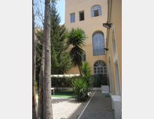 Cortile interno con ghiaia e vialetto in pietra, circondato da un edificio color crema con grandi finestre ad arco su più piani. Nello spazio verde sono presenti alberi alti e piante tropicali, con una zona ombreggiata da una copertura.