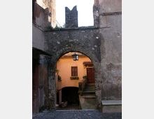 Arco in pietra di un vicolo medievale tra edifici intonacati, con una lanterna appesa al centro. Oltre l’arco si vede un cortile con scala in cemento che porta a una porta in legno e una finestra su una facciata color ocra.